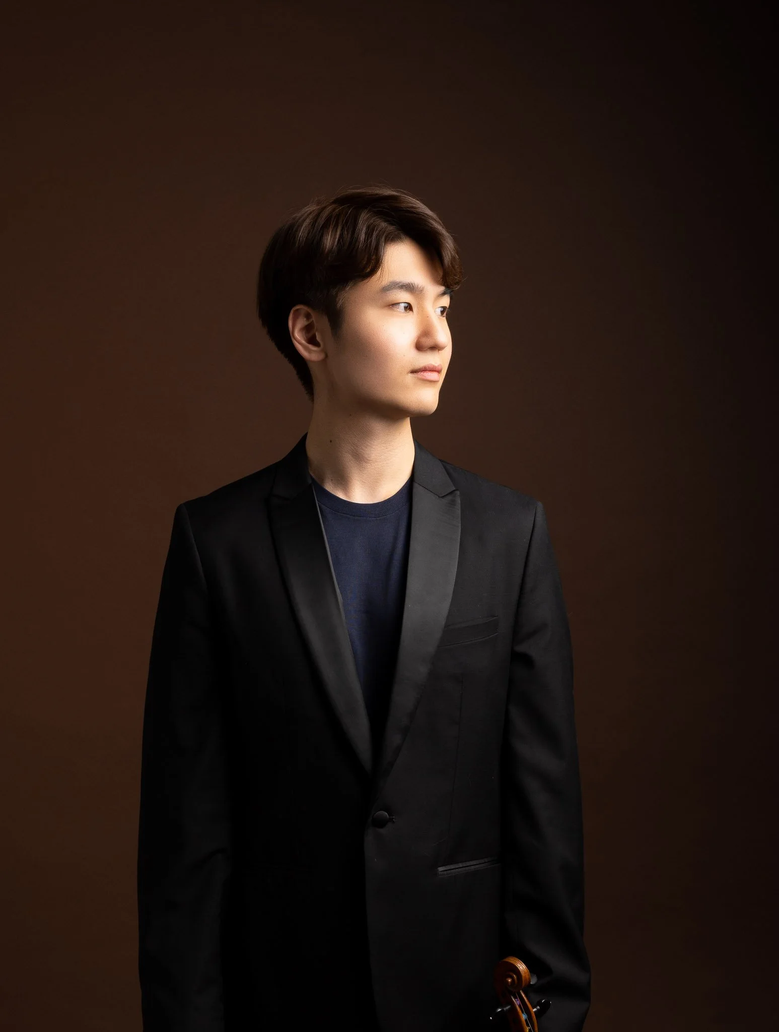Young man in a black tuxedo holding a violin and bow, looking to the right, with a brown gradient background.