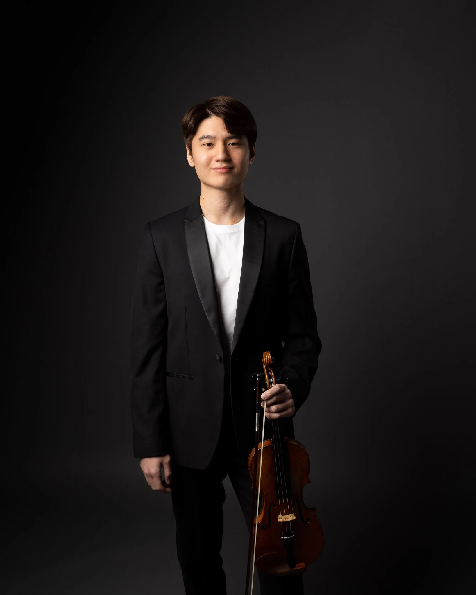 Young man in a black suit holding a violin against a dark background.