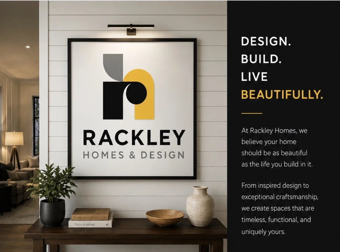 Interior of a home with a framed Rackley Homes & Design logo on a wall, next to a black panel with marketing text, a wooden table with decorative items, including a potted plant, books, a wooden bowl, and a ceramic vase.
