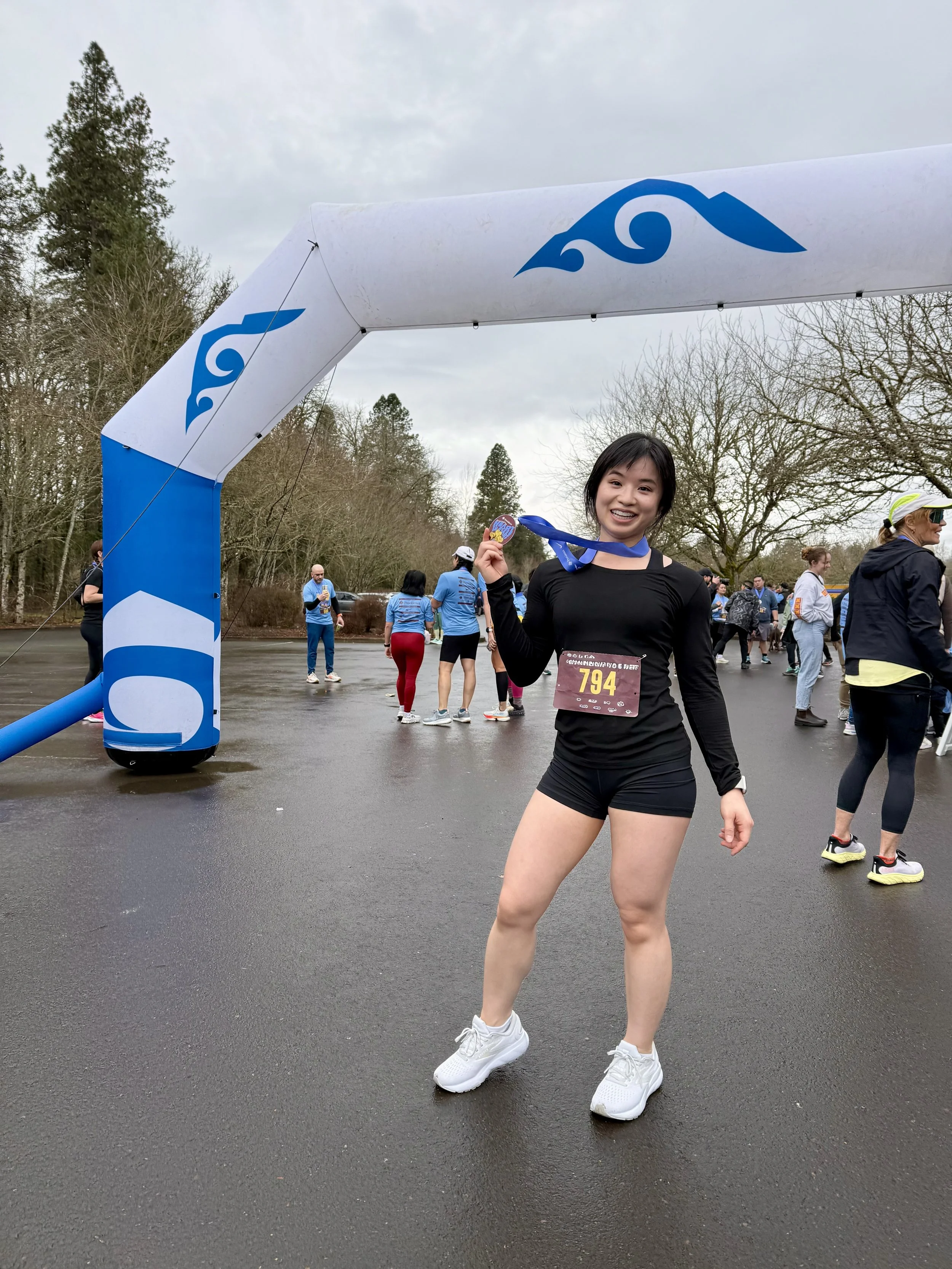 A young woman standing at a finish line event, holding her medal and smiling. She is dressed in a black long-sleeve shirt, black shorts, and white running shoes. There are several people in the background, some in athletic gear, with overcast weather and leafless trees surrounding the area.