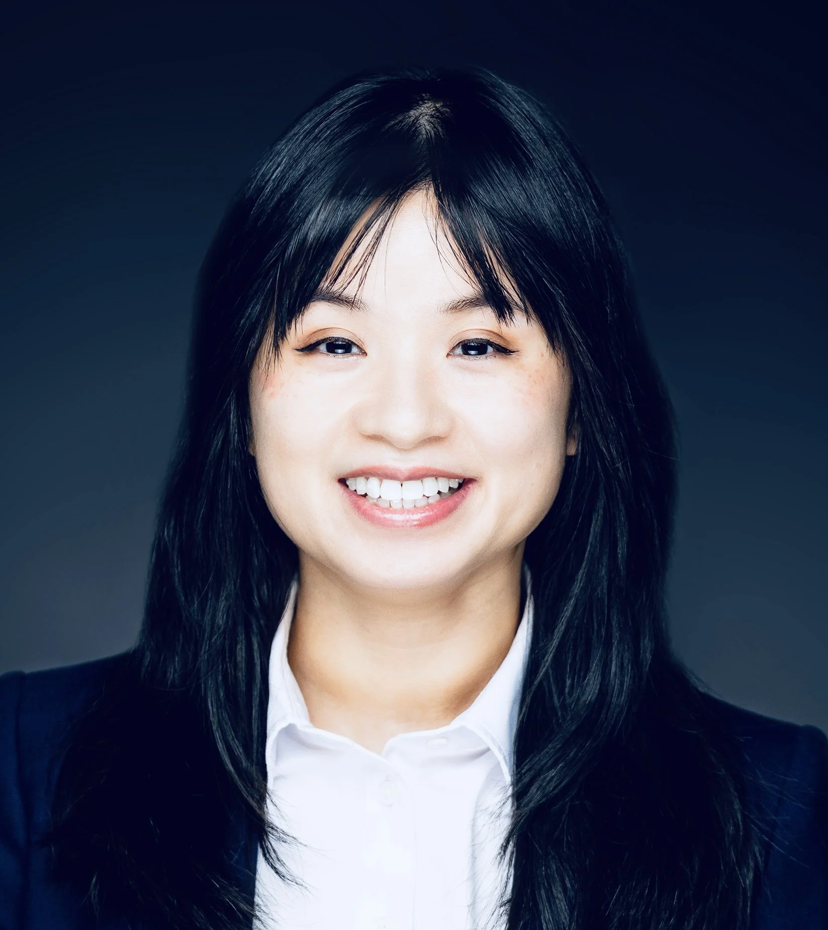 Headshot of a woman with black hair, smiling, wearing a dark blazer and white shirt, against a dark background.