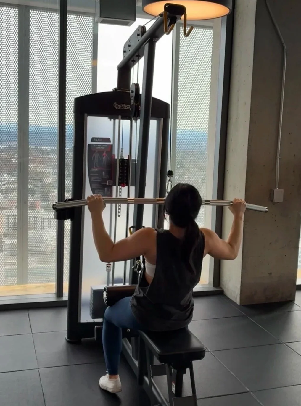 Woman using a seated shoulder press machine at a gym, facing a large window with a city view in the background.