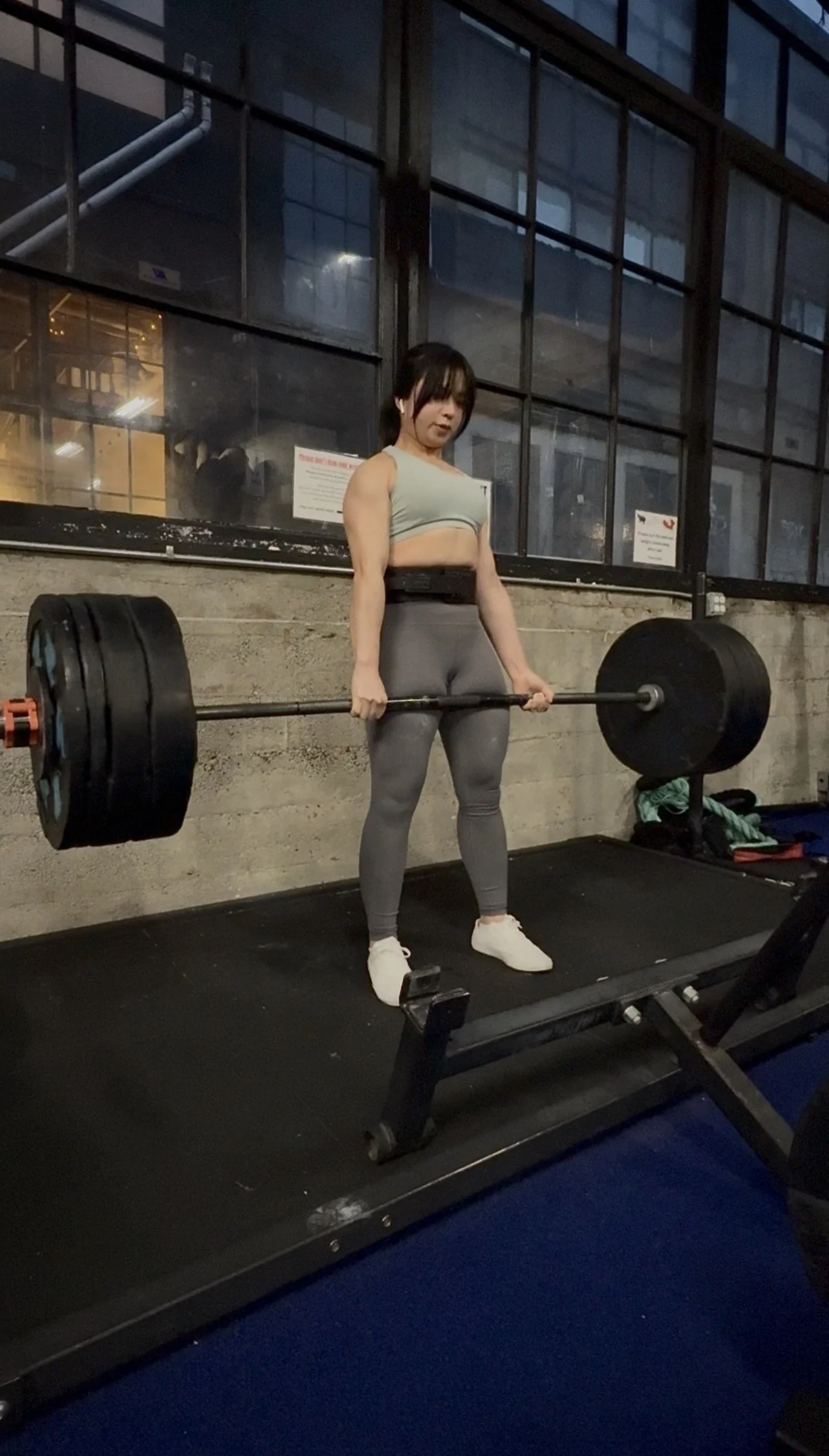 A woman wearing a sports bra and leggings is preparing to lift a heavy barbell at a gym. She is standing on a treadmill.