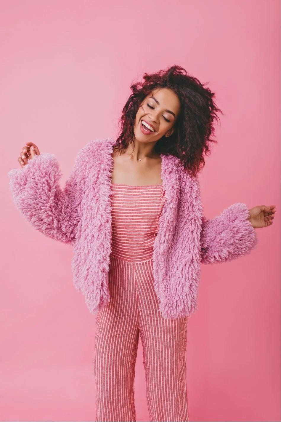 A woman with curly hair dancing against a pink background, wearing a pink furry jacket over a striped pink and white top and pink striped pants.