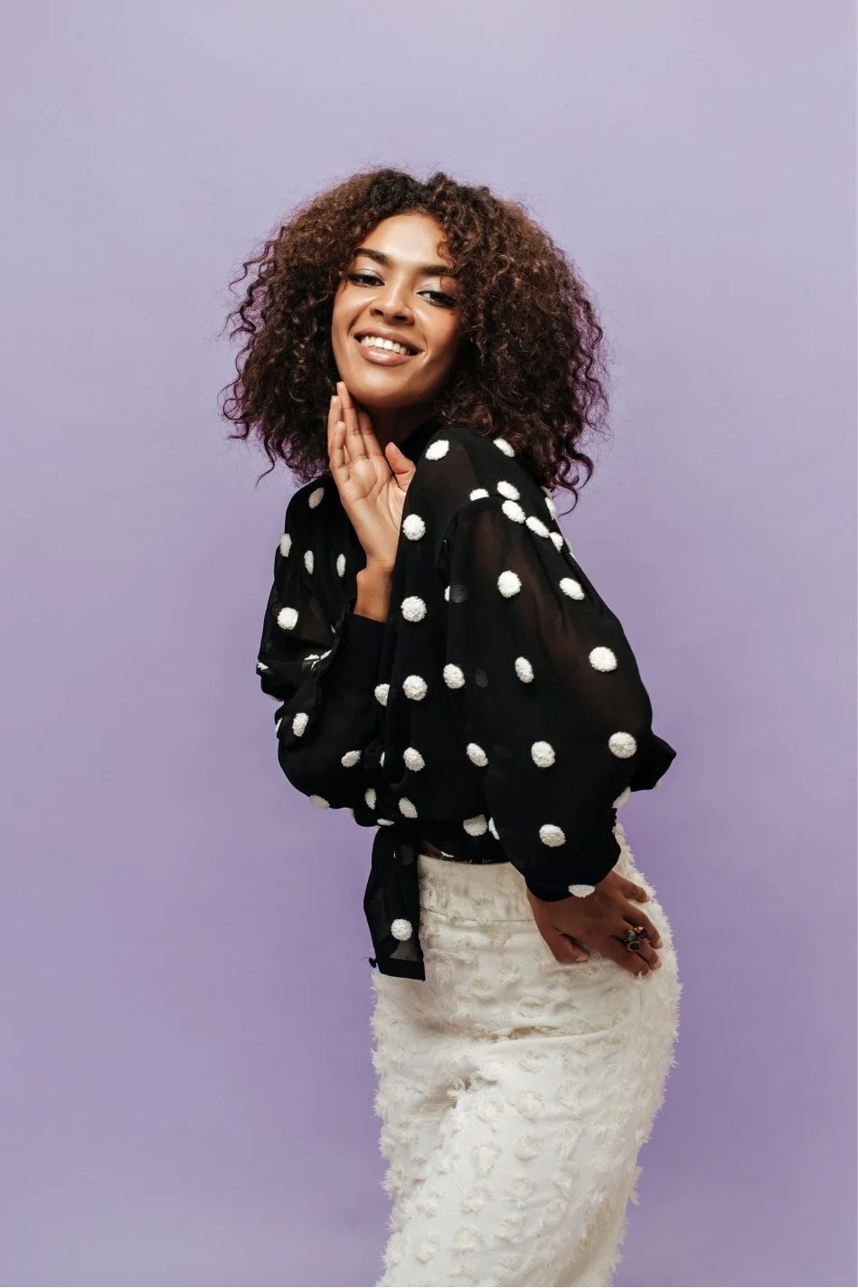 A woman with curly hair wearing a black blouse with white polka dots and a white textured skirt, smiling and posing against a purple background.