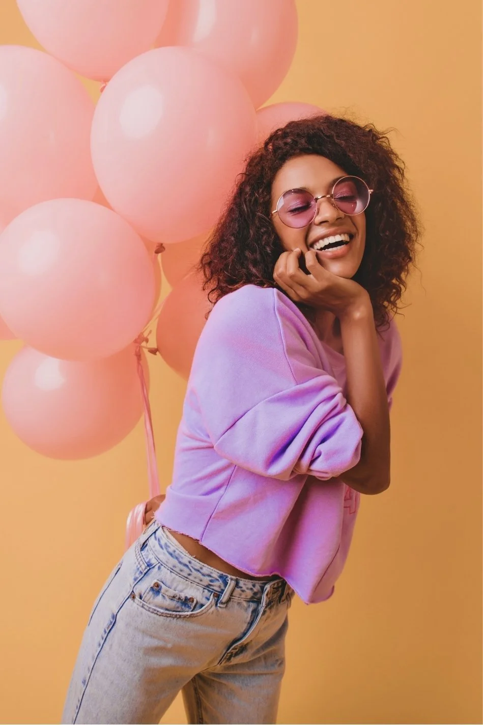A woman with curly hair wearing pink sunglasses and a pastel purple sweatshirt, smiling joyfully, standing in front of a background decorated with pink balloons and a yellow wall.