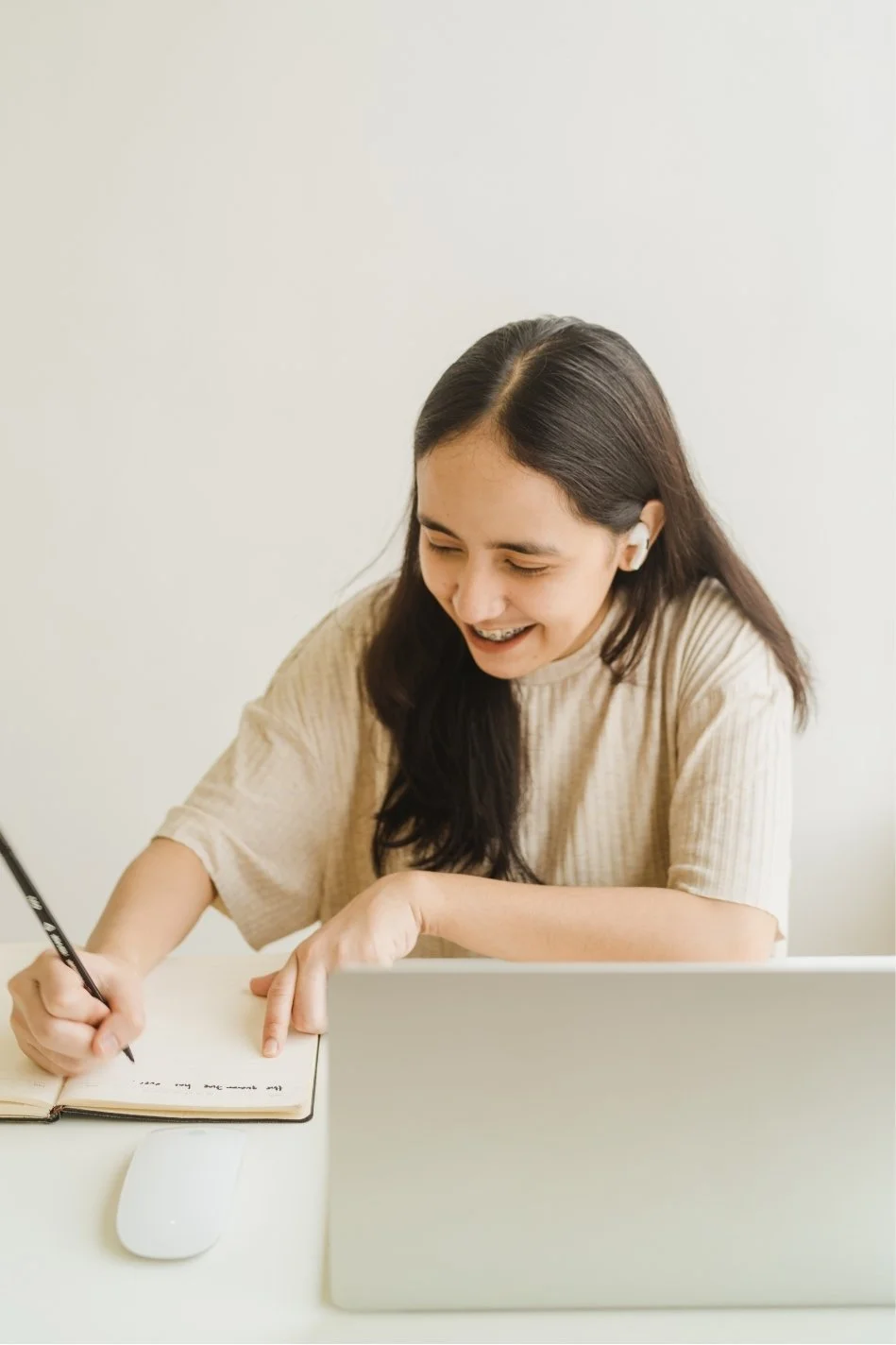 A young woman with long dark hair, smiling, sitting at a desk with a laptop, writing in a notebook, and wearing wireless earbuds.