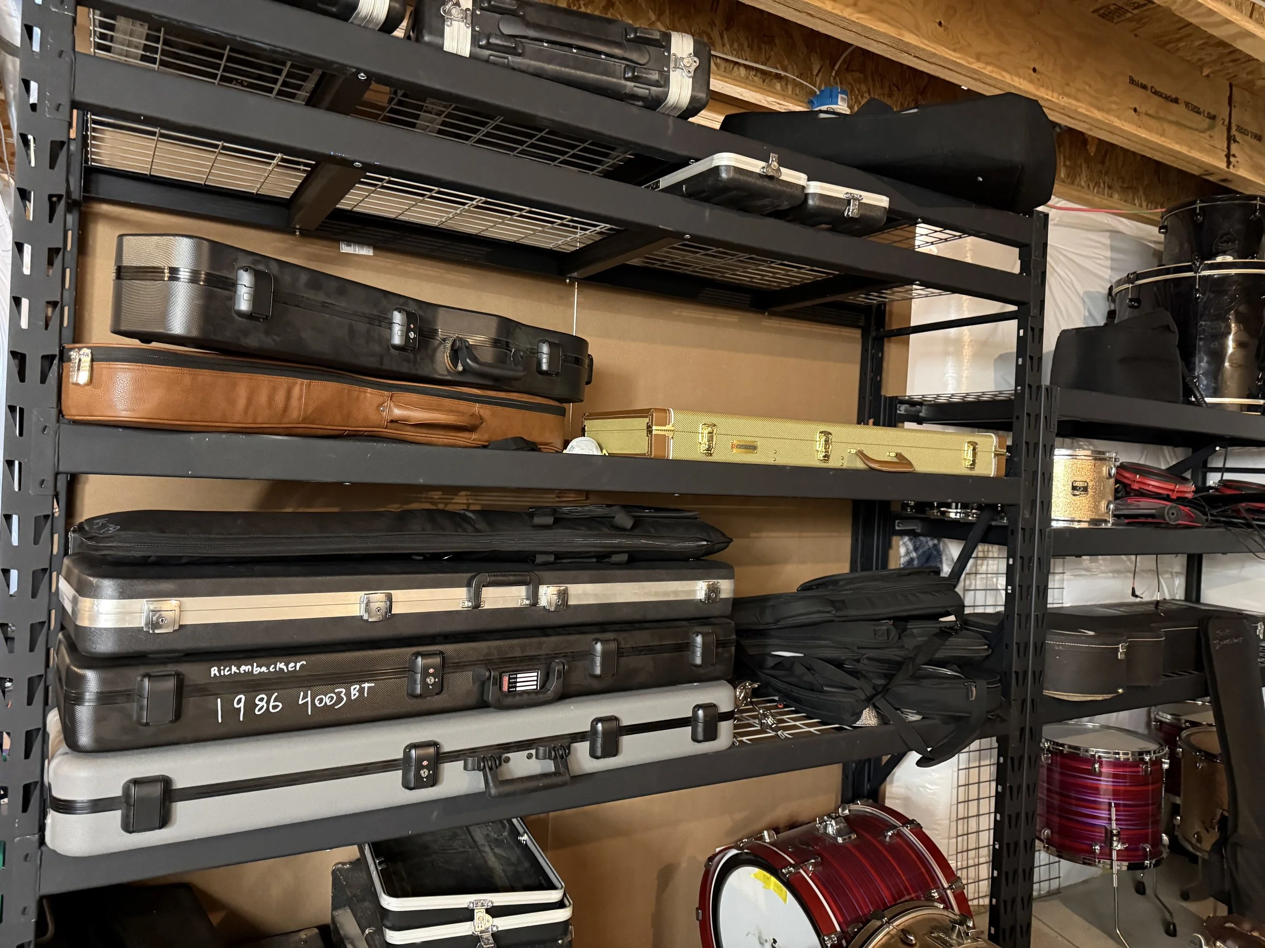 Black metal storage rack with several suitcases, a drum, and other musical equipment stored on it in a room with unfinished wood ceiling.