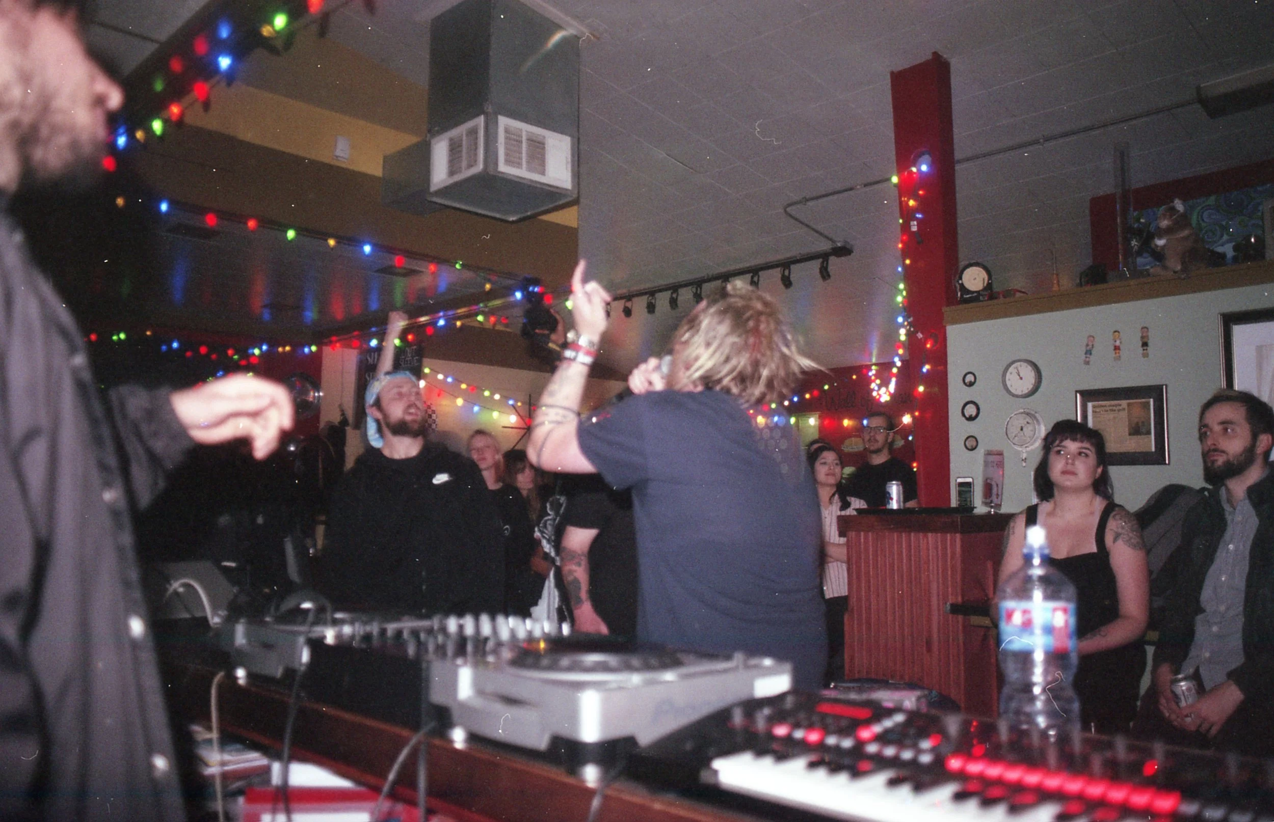 People dancing and socializing in a dimly lit room decorated with colorful string lights. A DJ or music equipment is visible in the foreground.