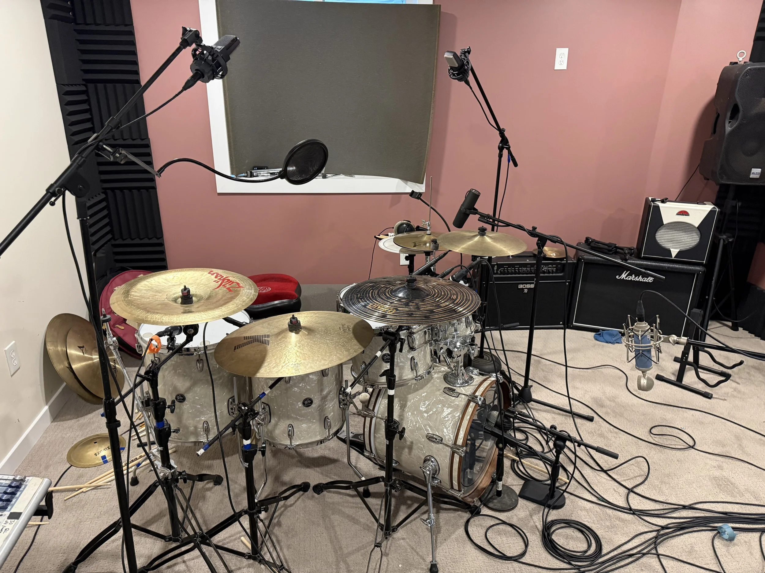 A music recording studio with drum set, microphones, speakers, and soundproofing. Pink walls and electrical outlets are visible.