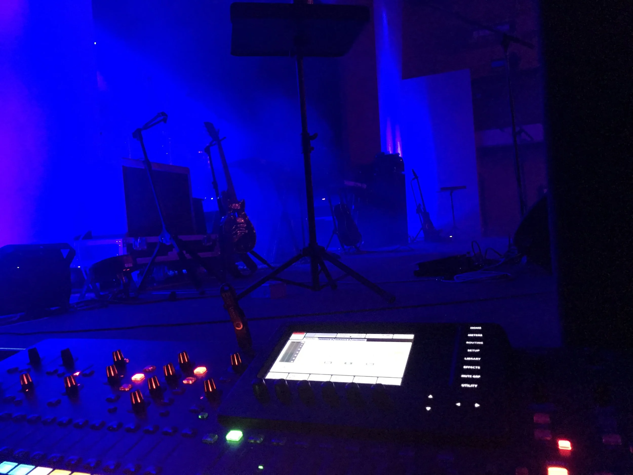 A stage with musical instruments, microphones, and speaker stands illuminated by blue and purple stage lights, with sound and music equipment in the foreground.