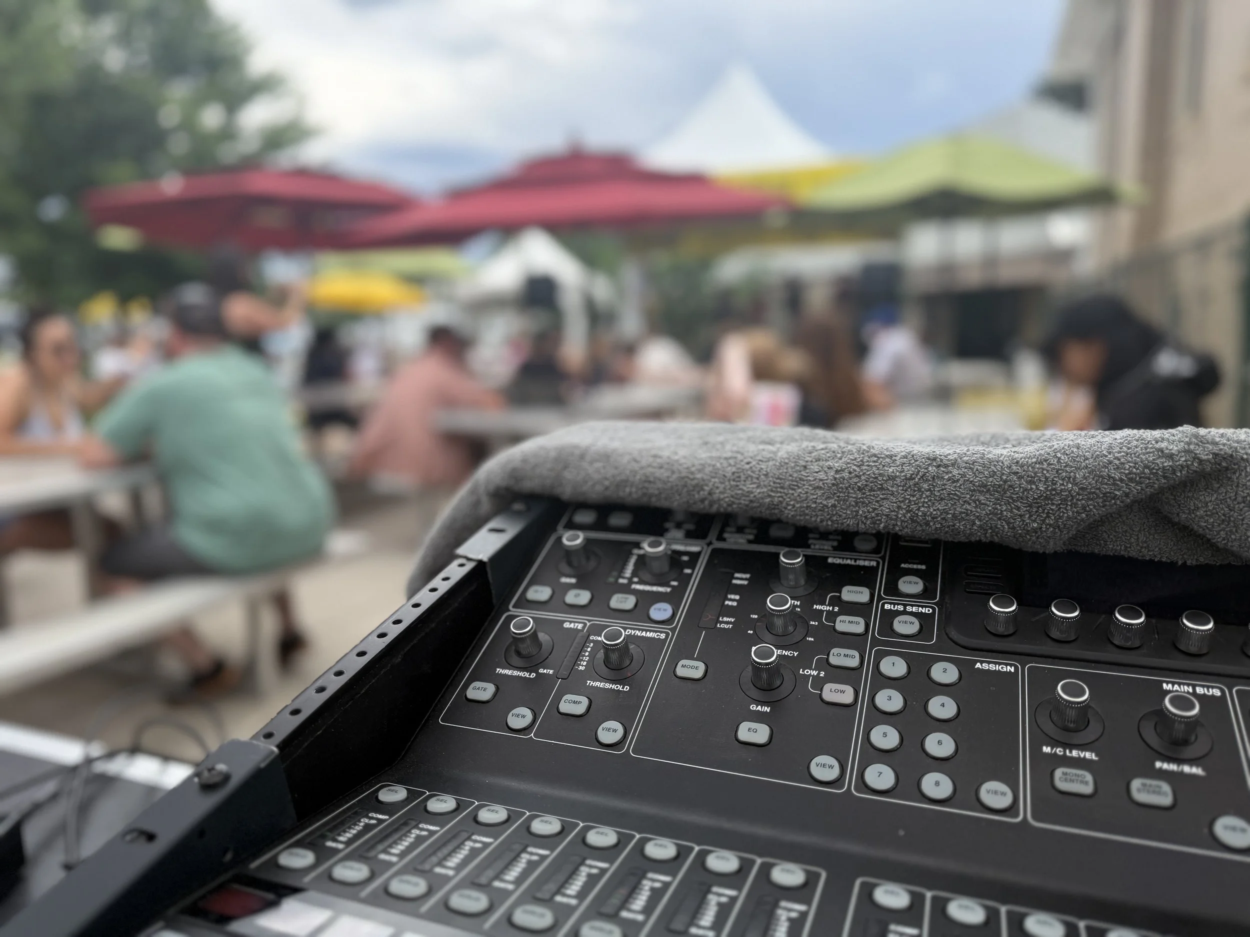 A DJ mixer with various knobs and sliders in the foreground, outdoor seating with red and yellow umbrellas, and people sitting at tables in the background, under a partly cloudy sky.