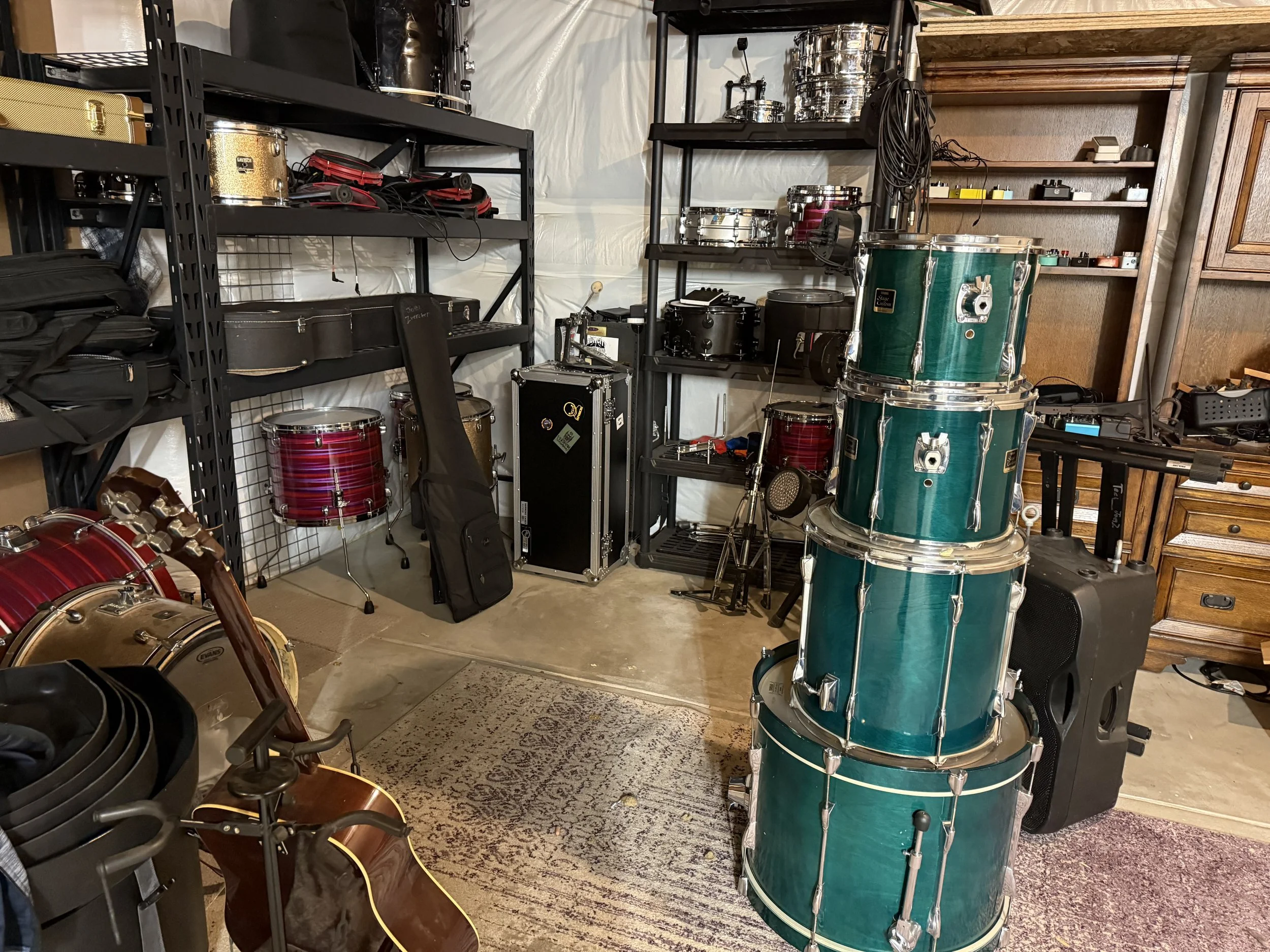 A collection of musical instruments including drums, a guitar, and various percussion equipment stored on black metal shelves and in a corner of a room.