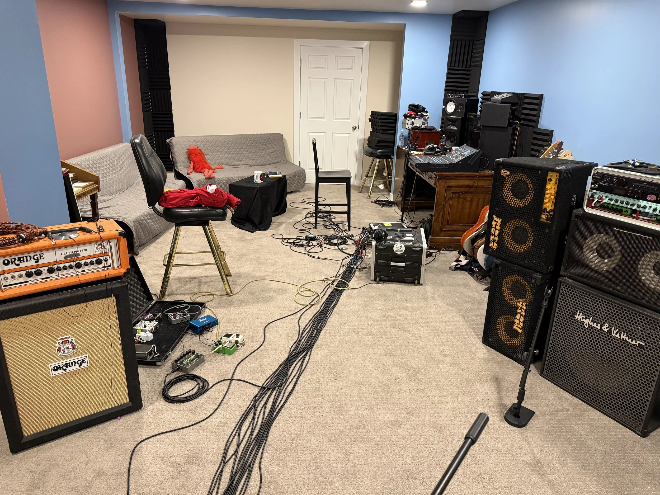 A music rehearsal or recording studio with guitars, amplifiers, speakers, and audio equipment set up. There are sofas, chairs, and a table with miscellaneous items, including a plush toy on one sofa. Cables are scattered across the floor.