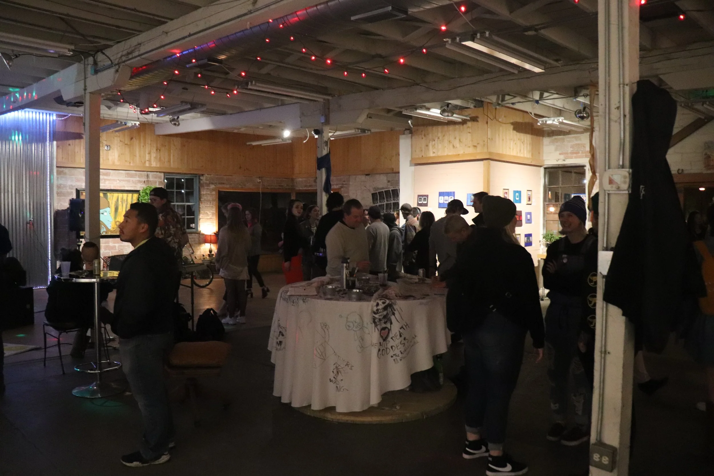 An indoor gathering with people socializing under string lights, with a table decorated with drawings and messages, in a cozy, rustic-style space with brick and wooden walls.