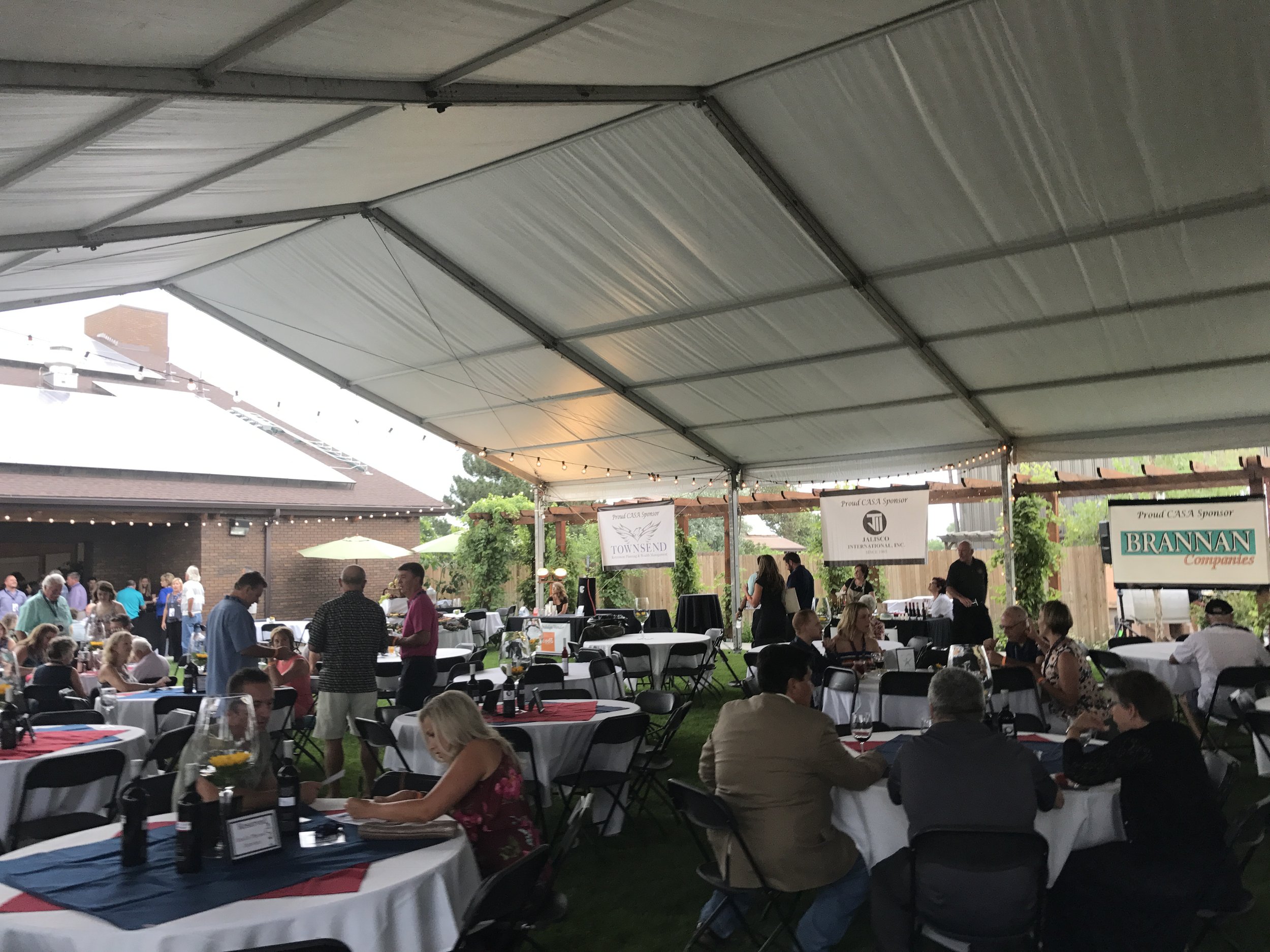 A large outdoor gathering under a metal canopy tent with people sitting at round tables and walking around. The tables are decorated with red, white, and blue tablecloths. There are banners hanging from the tent with various company logos. The background shows a brick building and some greenery.