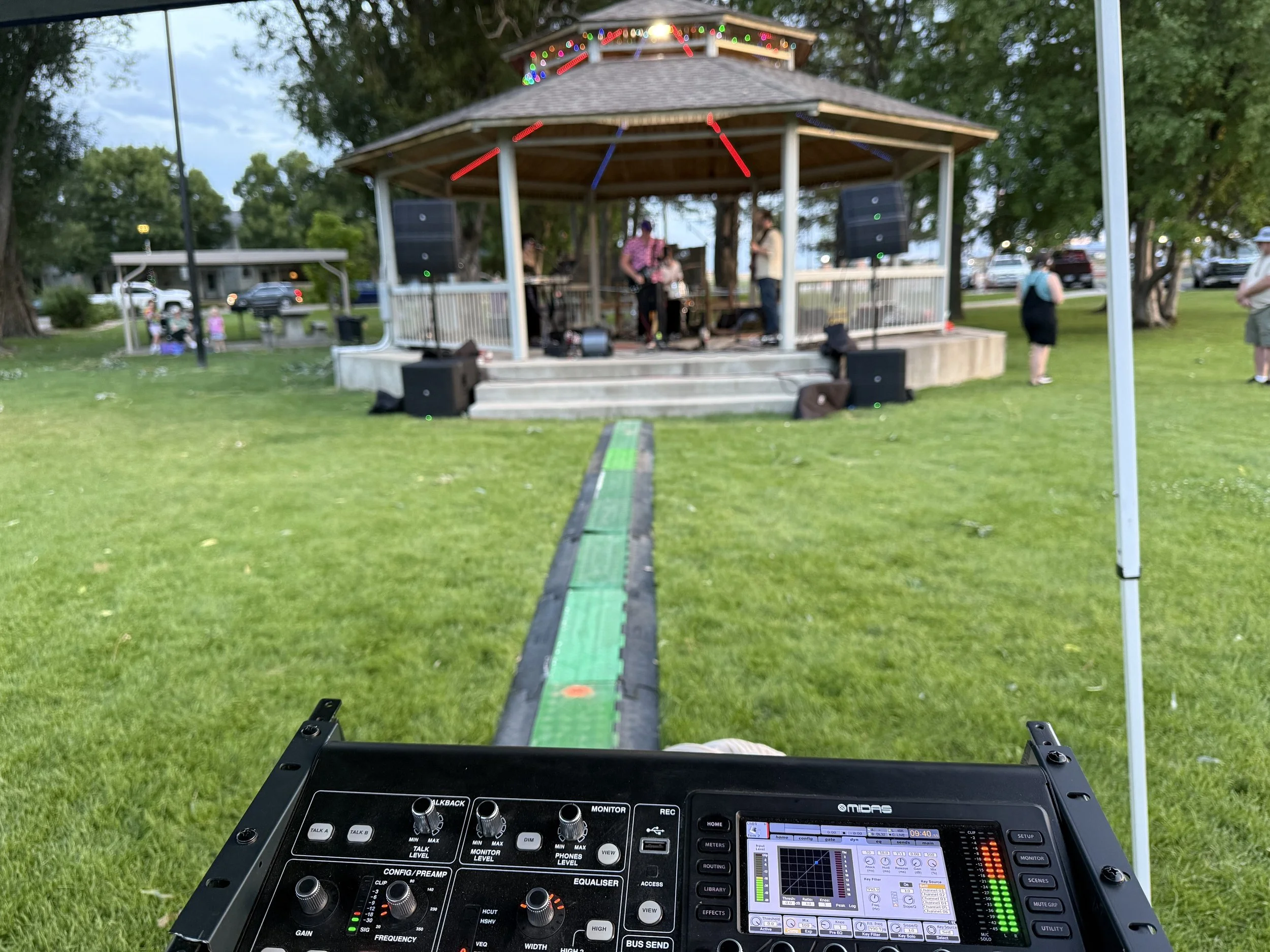 View from a DJ or sound engineer's perspective at an outdoor event, showing a sound mixing console in the foreground, a band performing on a gazebo stage in the background decorated with Christmas lights, surrounded by trees and people