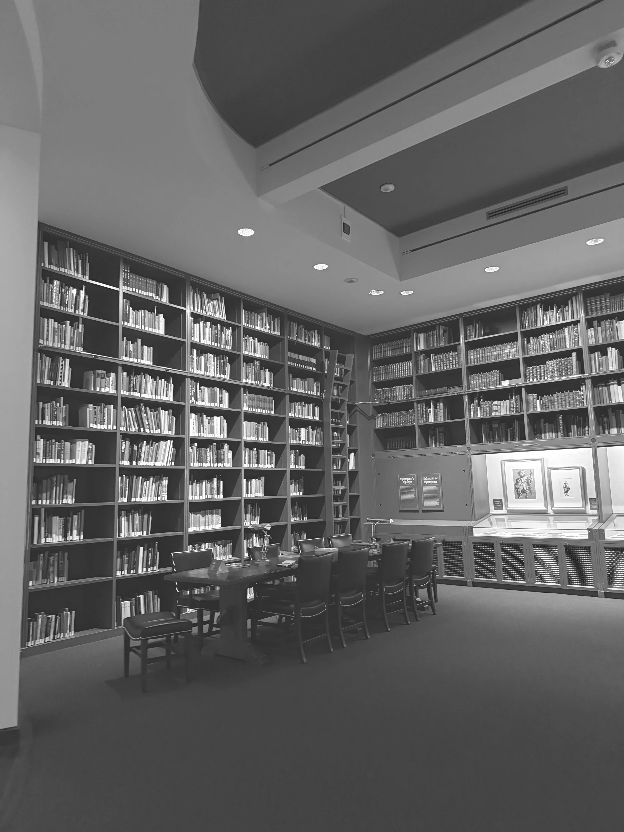 Black and white photo of a library reading room with tall bookshelves filled with books and a table surrounded by chairs in the foreground.