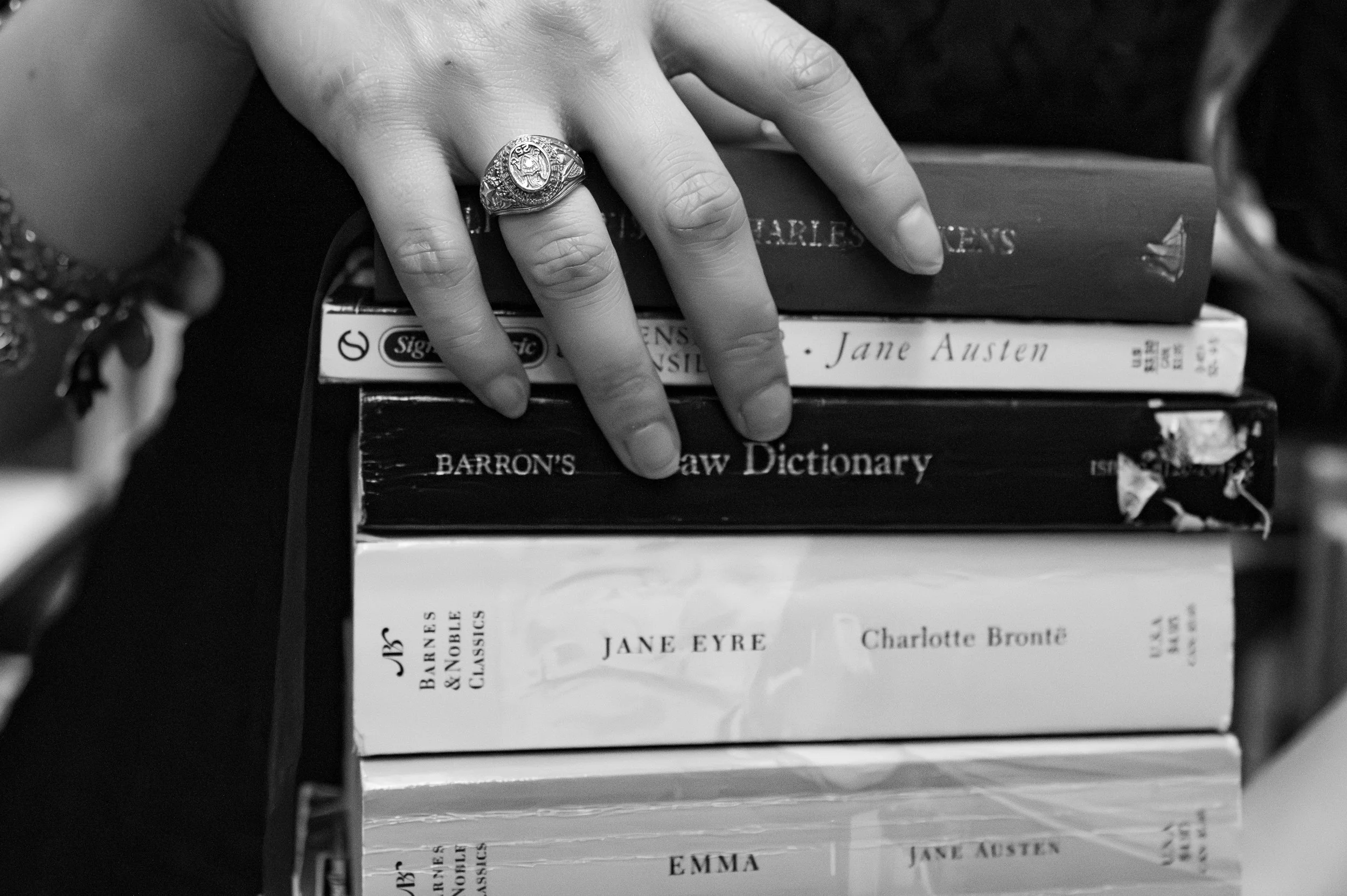 A close-up black-and-white photograph of a person's hand wearing a large block-style ring with a crest or emblem. The hand is resting on a stack of books, including titles by Jane Austen like "Emma" and "Persuasion" with visible authors' names. The scene appears to be indoors, possibly on a bookshelf or desk.