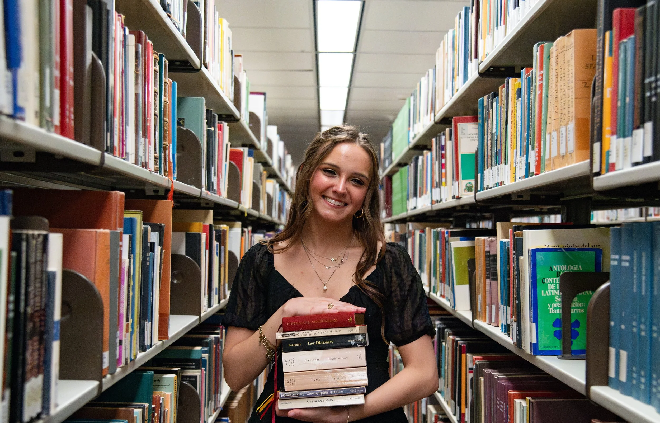 A young woman with light brown hair smiling at the camera, standing in a library between bookshelves, holding a stack of books.