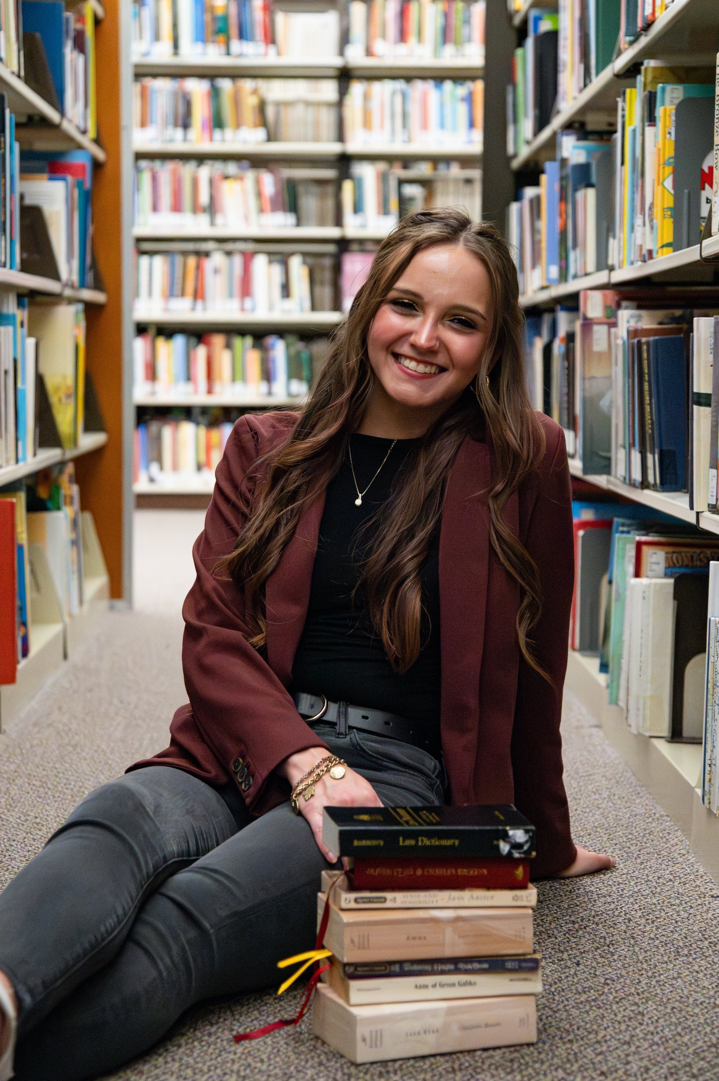 A young woman with long brown hair smiling while sitting on the floor of a library, surrounded by shelves of books. She has a stack of books in front of her, including a dictionary, and is wearing a maroon blazer, black top, and jewelry.