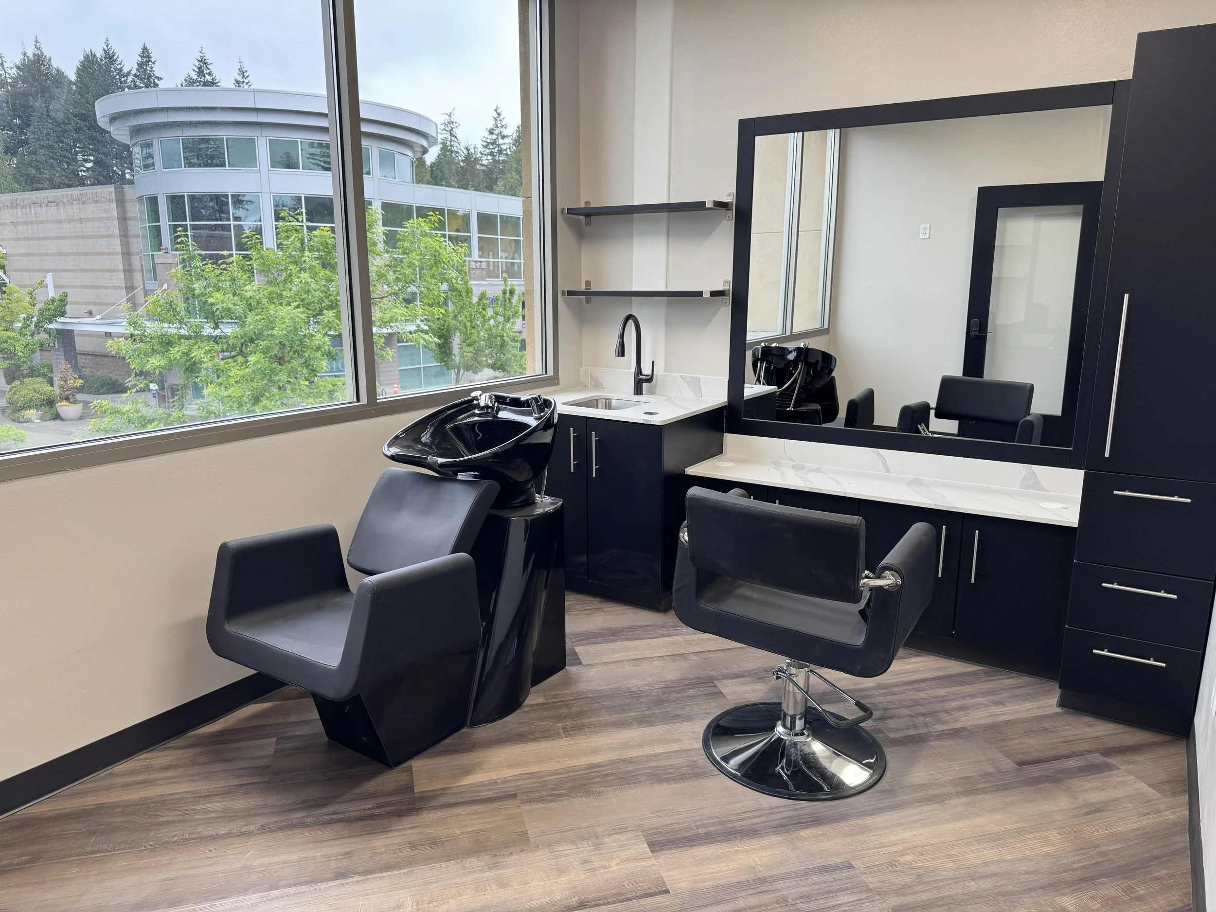 Interior of a hair salon or barbershop with black styling chairs, a sink, and large windows showing trees and a modern building outside.