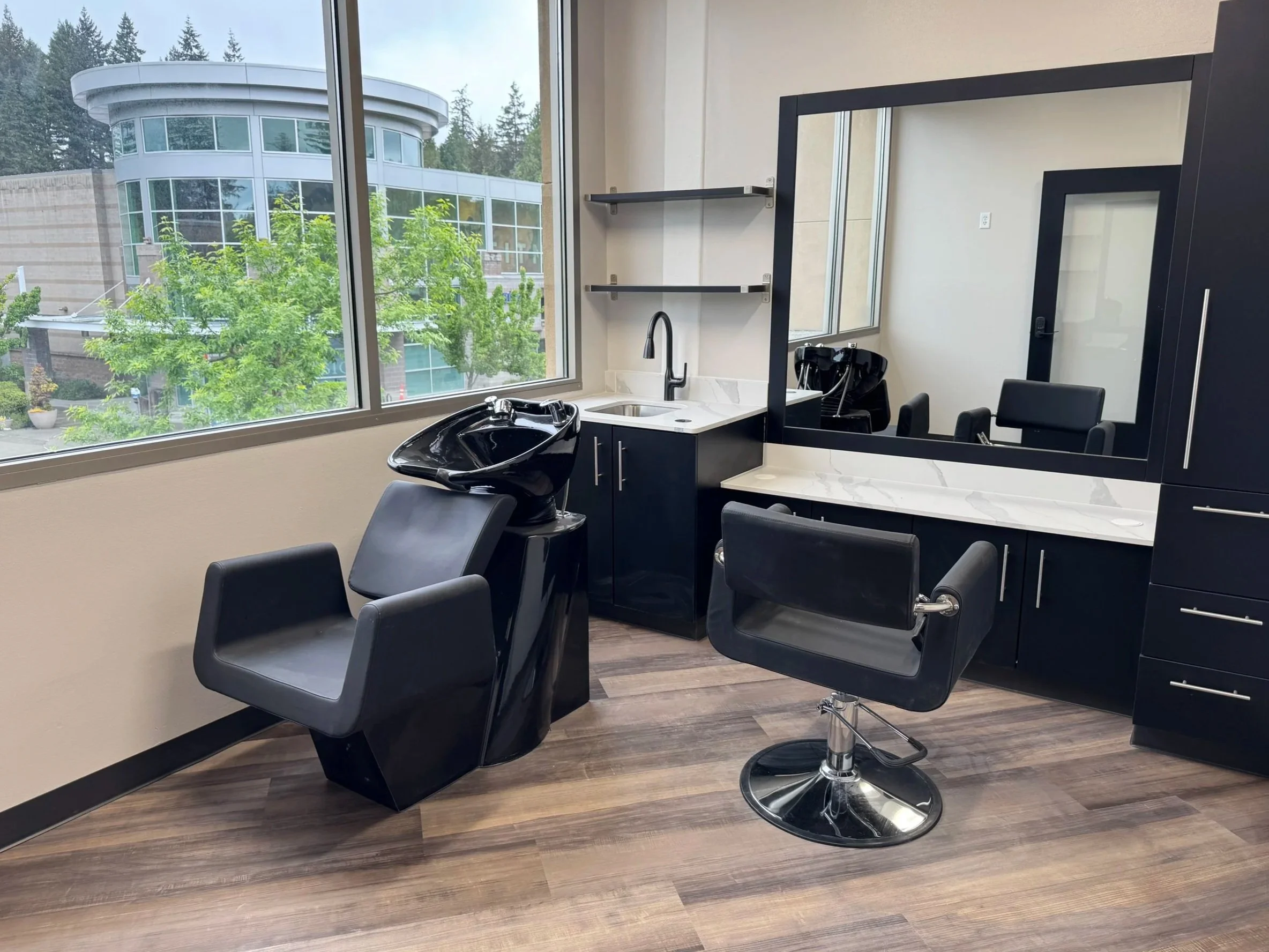 Interior view of a hair salon with black styling chairs, a hair wash sink, and a large window showing a building and trees outside.