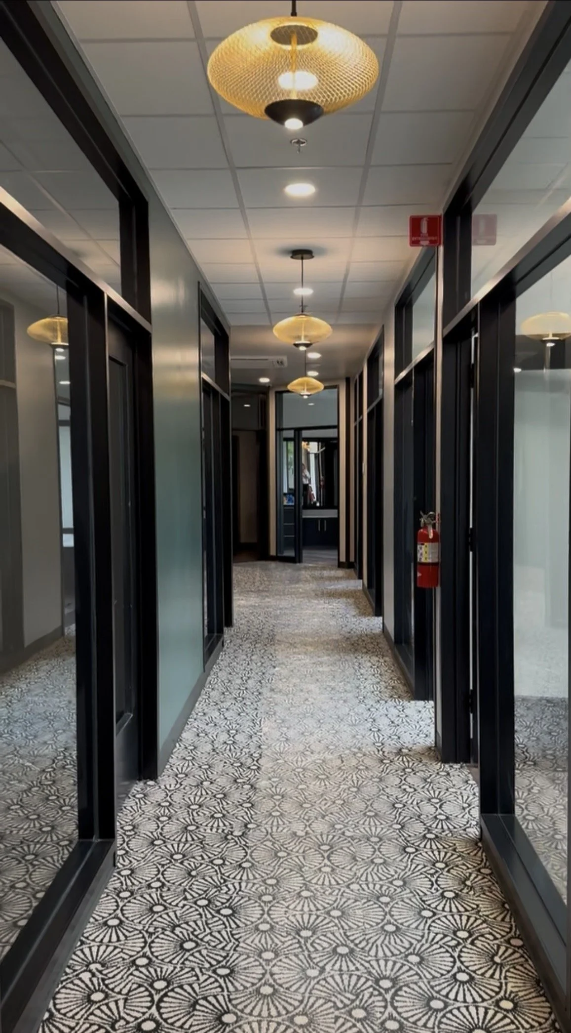 Interior of a hallway with patterned tile flooring, black-framed glass doors on each side, and yellow ceiling lights. Fire extinguisher is mounted on the right wall.