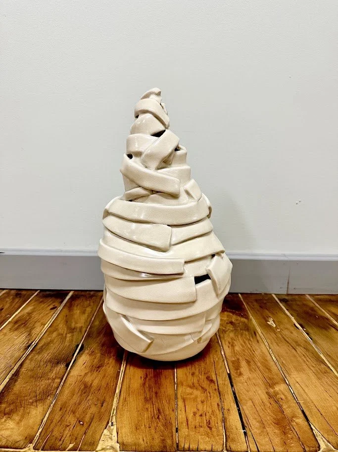 A ceramic sculpture resembling a stack of irregularly shaped, creamy white blocks, arranged in a tapered, conical form. The sculpture is placed on a wooden floor against a plain, light-colored wall.