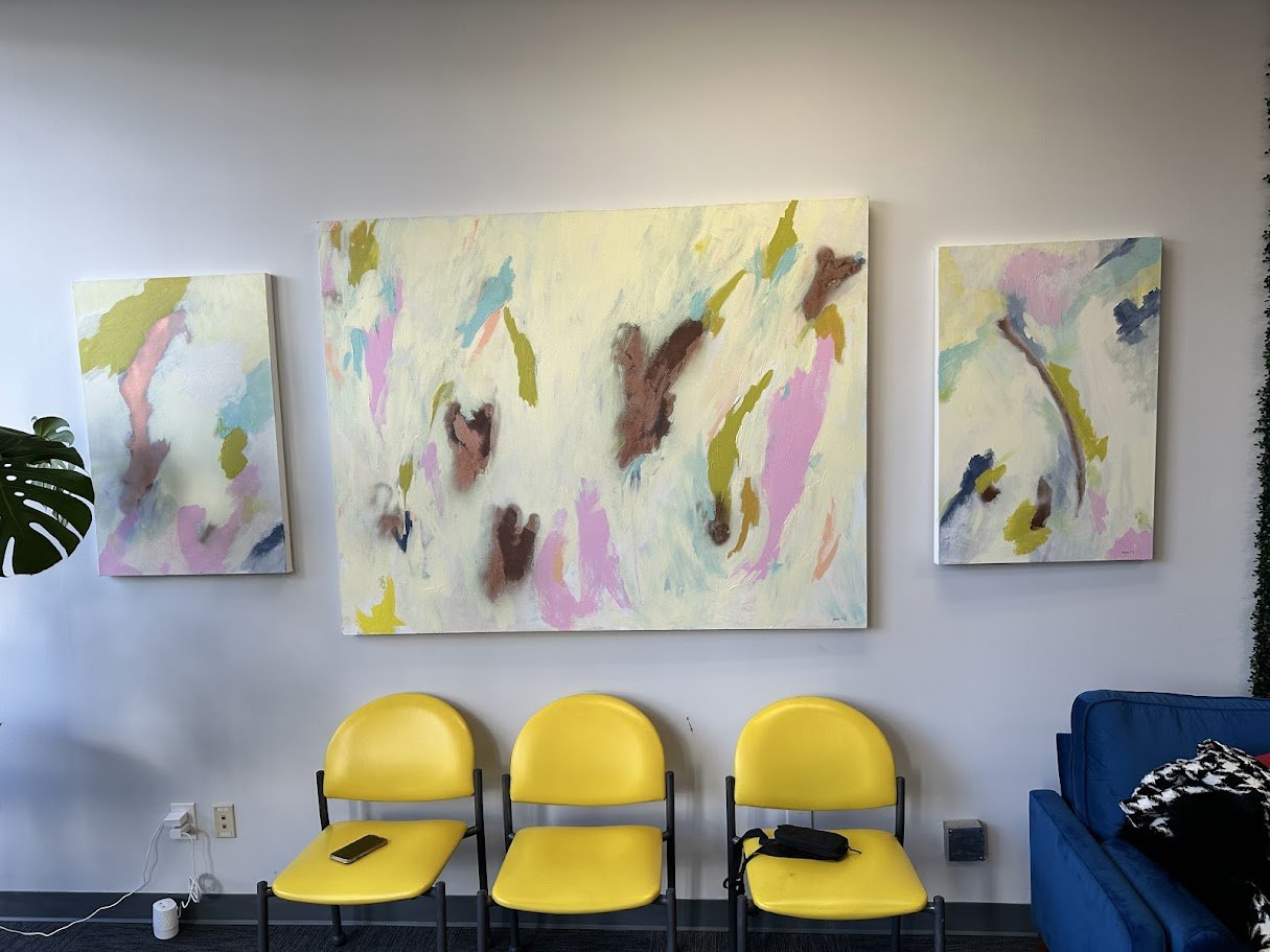Three yellow chairs arranged in front of a white wall with abstract paintings, and a blue sofa on the right side of the image.