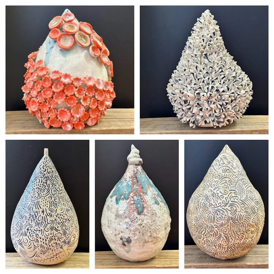 Five ceramic sculptures, three teardrop-shaped and two with rounded, irregular shapes, with intricate patterns and textures, displayed on a wooden surface against a black background.