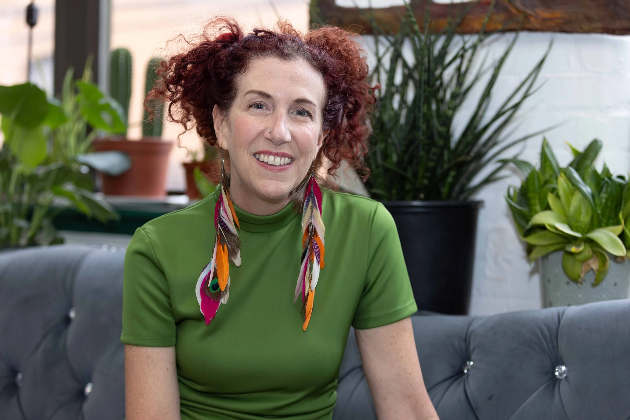 A woman with curly red hair wearing a green shirt and colorful feather earrings, smiling and seated on a black leather couch with green plants in the background.