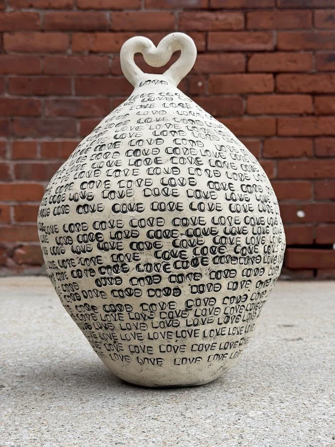 Decorative ceramic vase with the word 'love' repeatedly printed in black, featuring a heart-shaped handle, placed on a concrete surface against a brick wall.