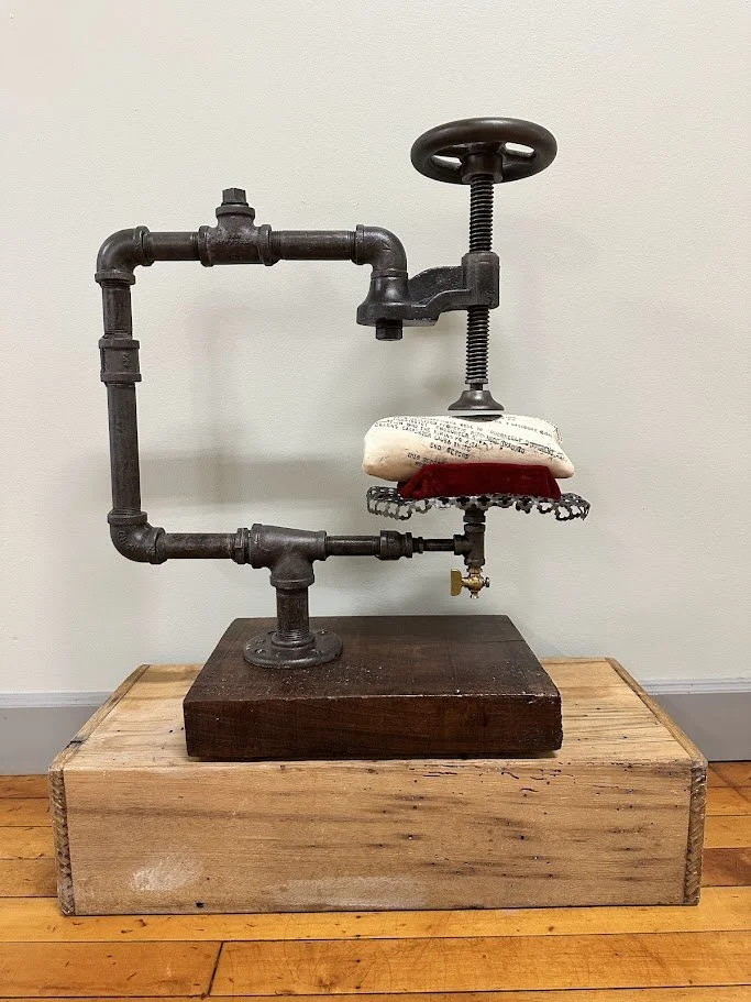 An artistic sculpture made from industrial pipes, a white pillow, a red object, and a small chain, mounted on a wooden base and displayed on a wooden surface.