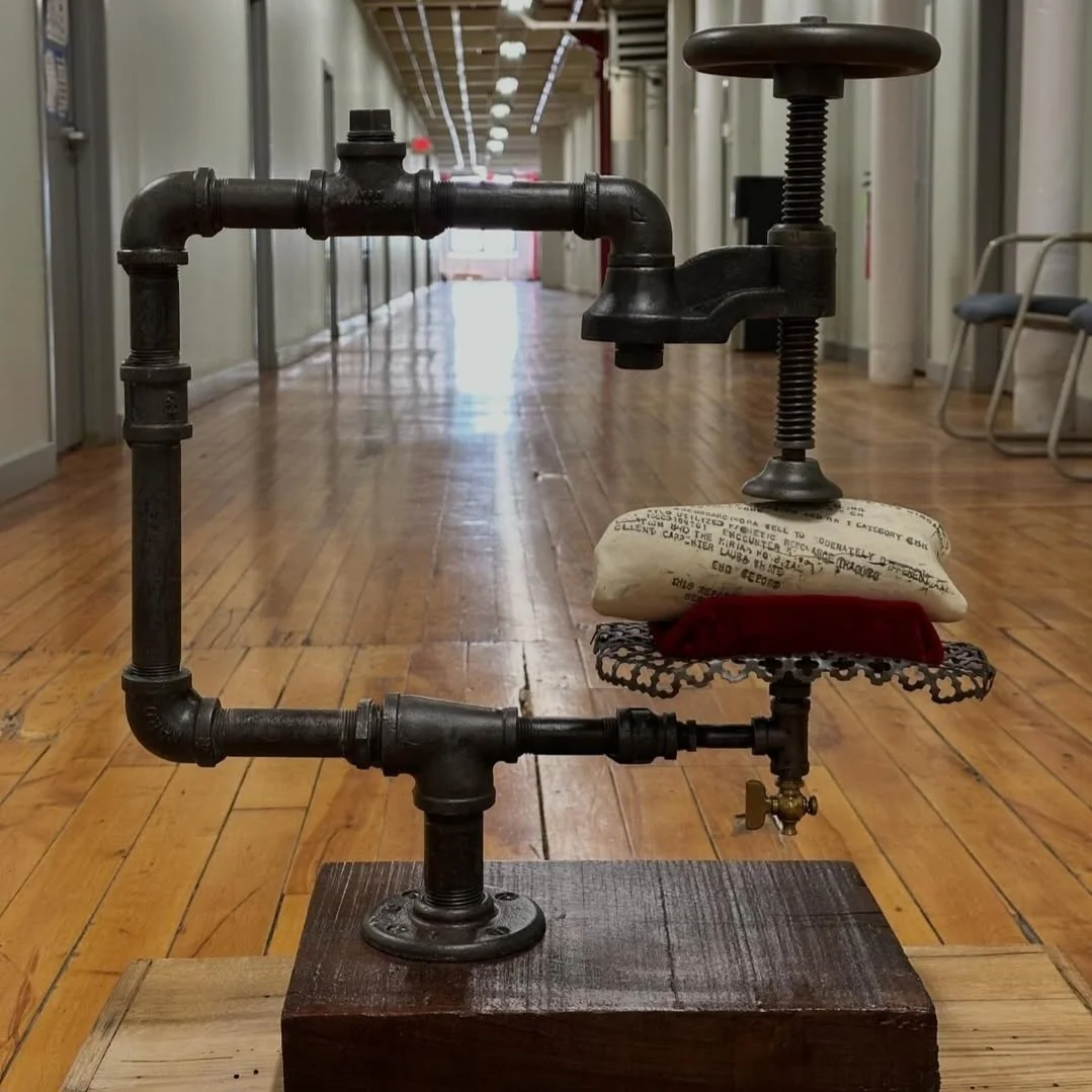 An artistic sculpture made of black iron pipes, clamps, and valves, resembling a desk with pillows on top, placed on a wooden base in a hallway with a wooden floor and chairs in the background.