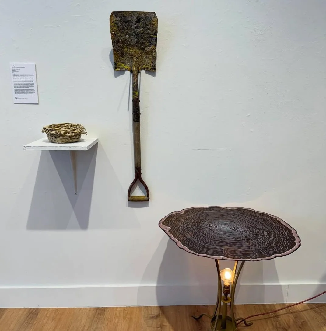 A wooden table with a natural, irregular edge, a light bulb attached beneath, and three slender legs. On the wall, there is an old rusted shovel and a basket on a small white shelf with a sticker.