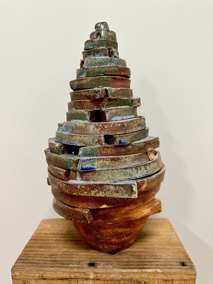 A ceramic sculpture resembling a layered cone or volcano placed on a wooden base against a plain background.