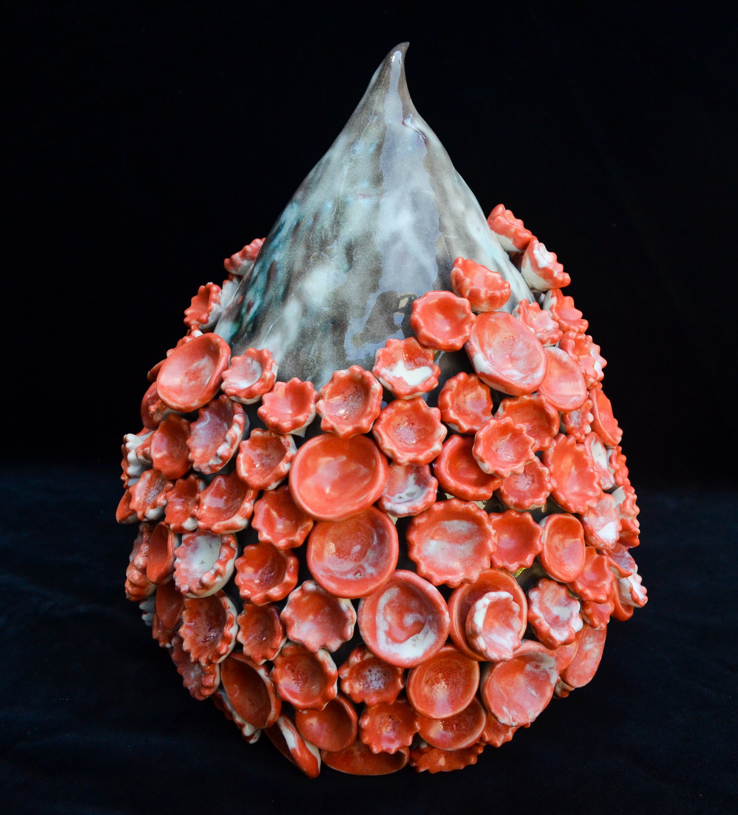 A geometric sculpture made of seashells and other materials, with a pointed top and surrounded by red shells, set against a black background.