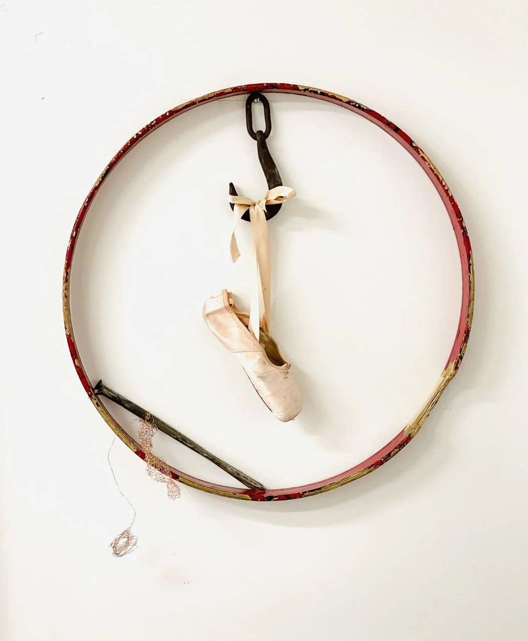 A clock frame with ballet pointe shoes hanging from the center, tied with a ribbon, and a ballet ribbon and brush hanging from the bottom part of the clock frame.
