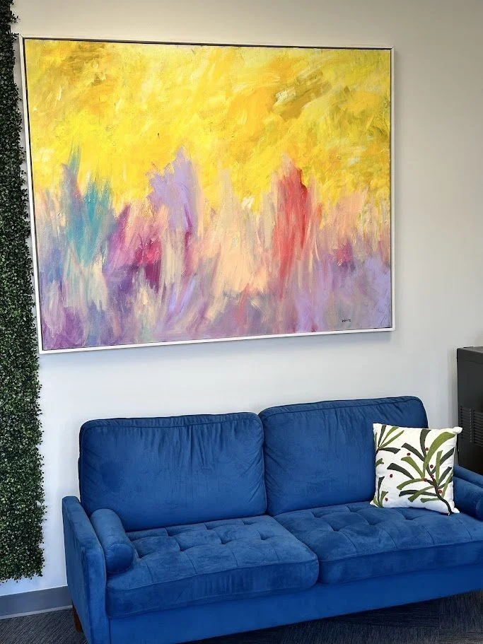 An abstract painting with yellow, purple, pink, and blue brushstrokes hanging above a blue couch with a decorative pillow featuring green and black leaf patterns.