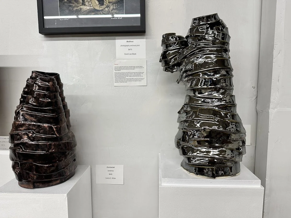 Two large ceramic sculptures resembling stacked, organic-shaped forms in dark, glossy finishes displayed on white pedestals in an art gallery.
