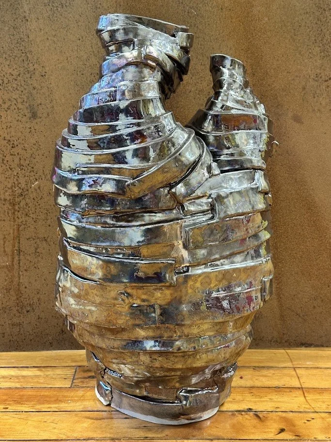 A metal sculpture resembling a human torso, made from layered, shiny, irregularly shaped metal pieces, placed on a wooden surface against a brown background.