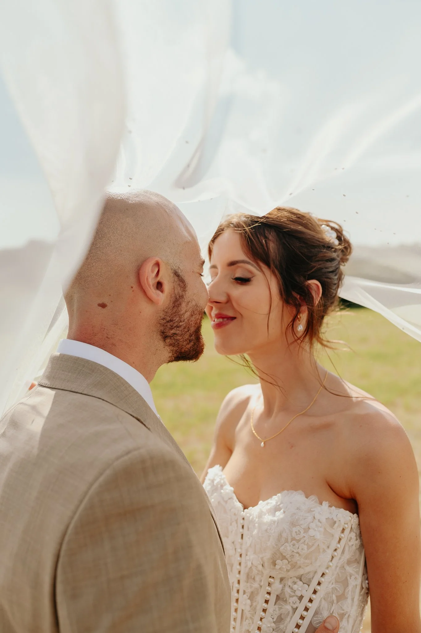A newlywed couple is sharing a close and intimate moment outdoors, with their noses almost touching. The groom is wearing a light-colored suit, and the bride is dressed in a white lace wedding gown with floral details and pearl accents. The bride has red hair styled in an updo and is wearing pearl earrings and a gold necklace. The background shows a bright, clear sky and a grassy field.