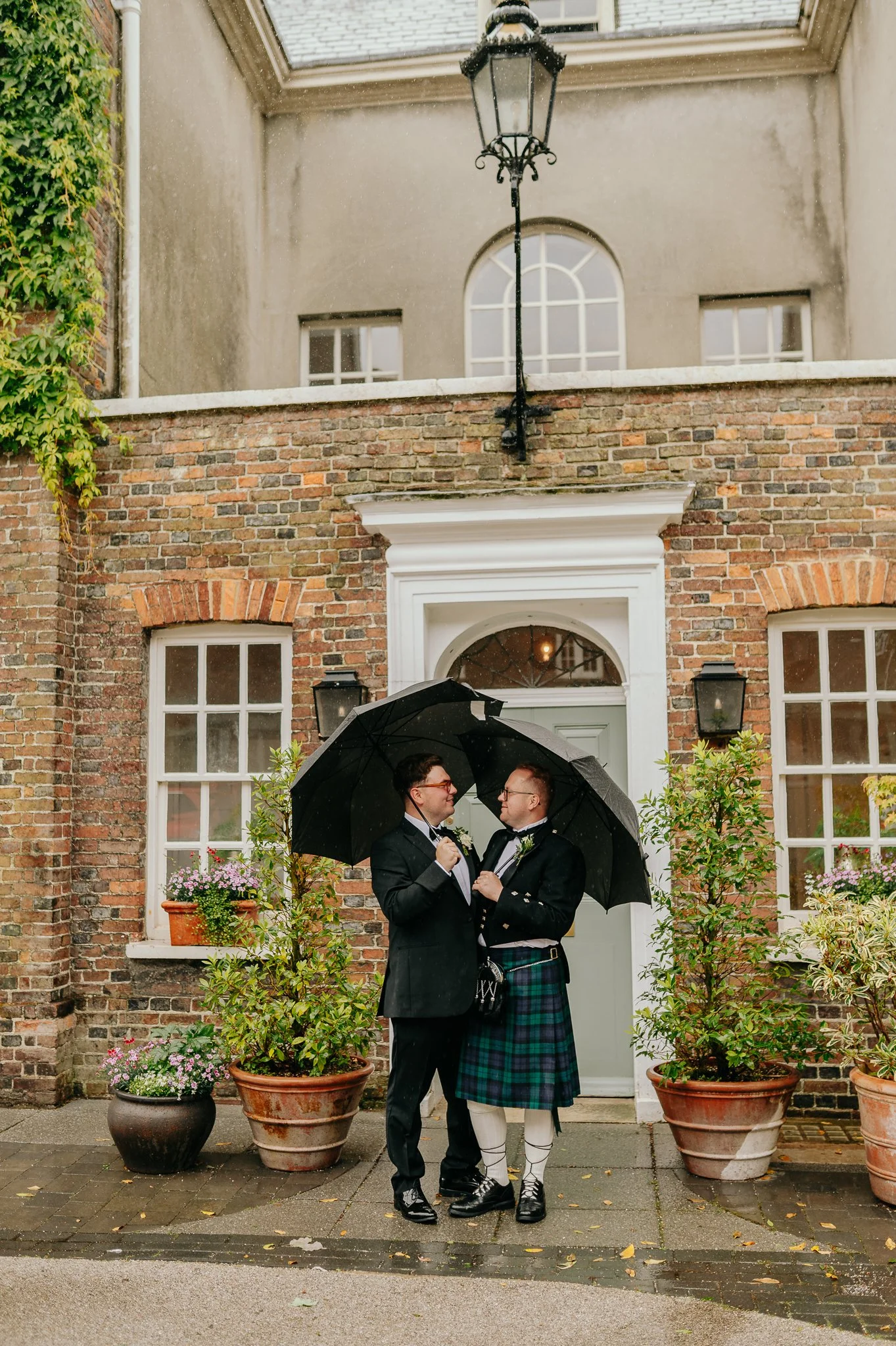 Groom and groom smile happily at each other holding black umbrellas outside wedding venue Pelham House in the rain