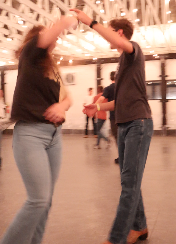 A young man and woman dancing together in a lively, indoor setting with string lights overhead.