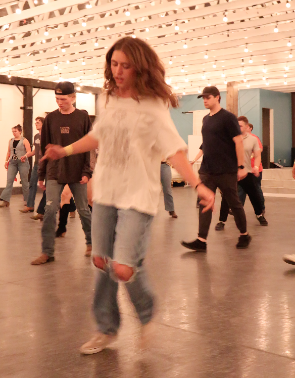 Group of people line dancing in a spacious hall with string lights on the ceiling, a woman in the foreground wearing ripped jeans and a loose white shirt leading the dance.