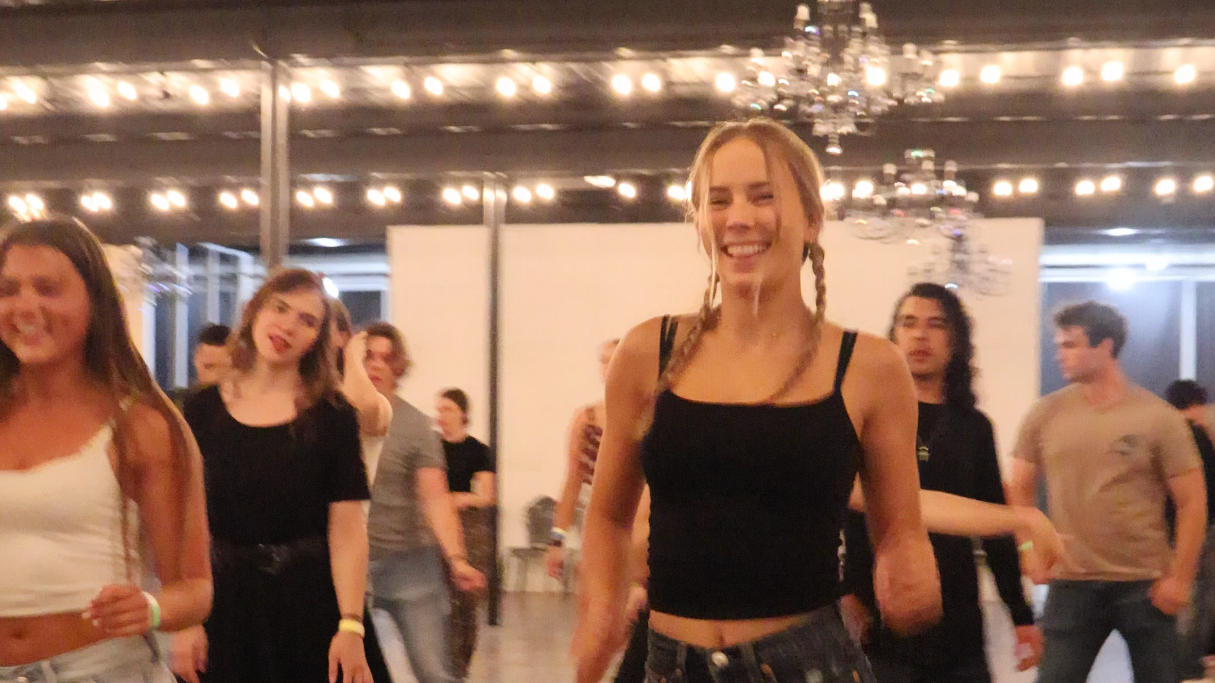 Young woman with braided hair smiling at Marnell's Country Dance Iron Elegance doing a line dance, surrounded by other people in a decorated indoor setting with string lights and a chandelier.