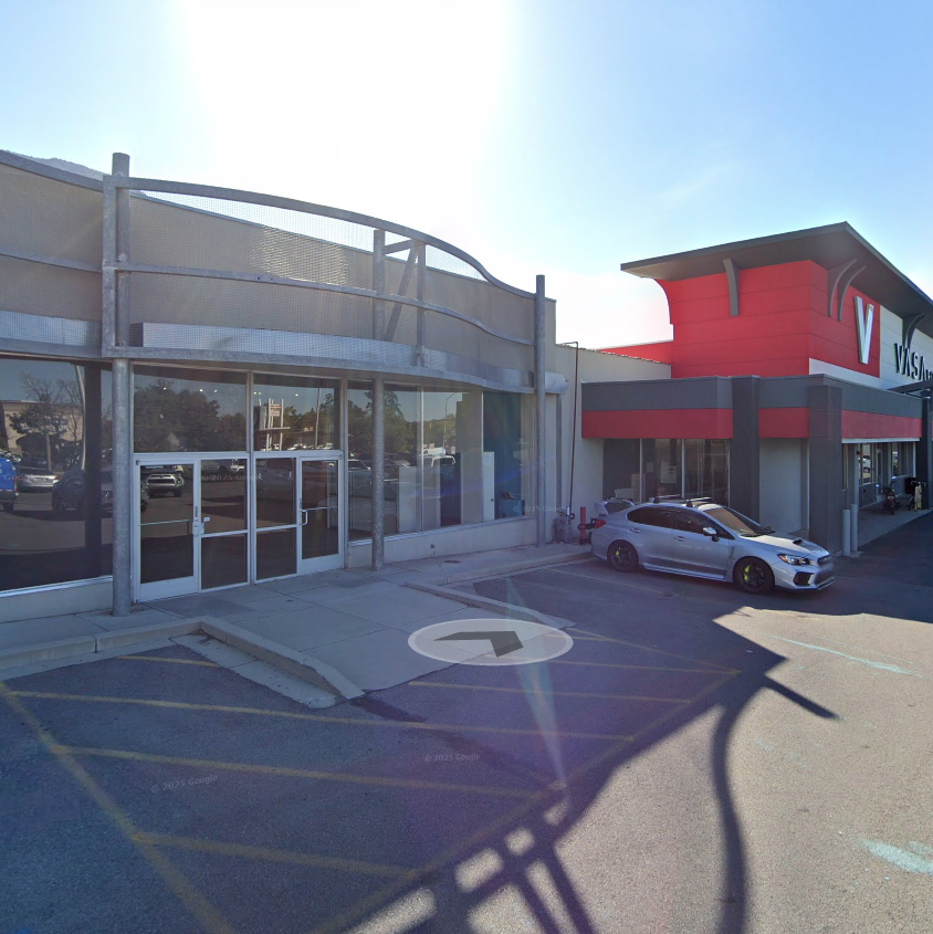 Outside of Iron Elegance Venue next to Vasa Fitness in Provo with a parking lot and a silver car parked near the entrance. Clear sky with bright sunlight.