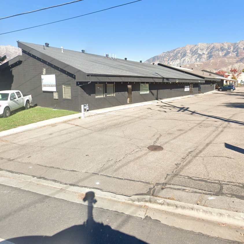 Rustic Paradise venue in Orem with a sloped gray roof, located in a parking lot with a few parked cars and a mountain range in the background.