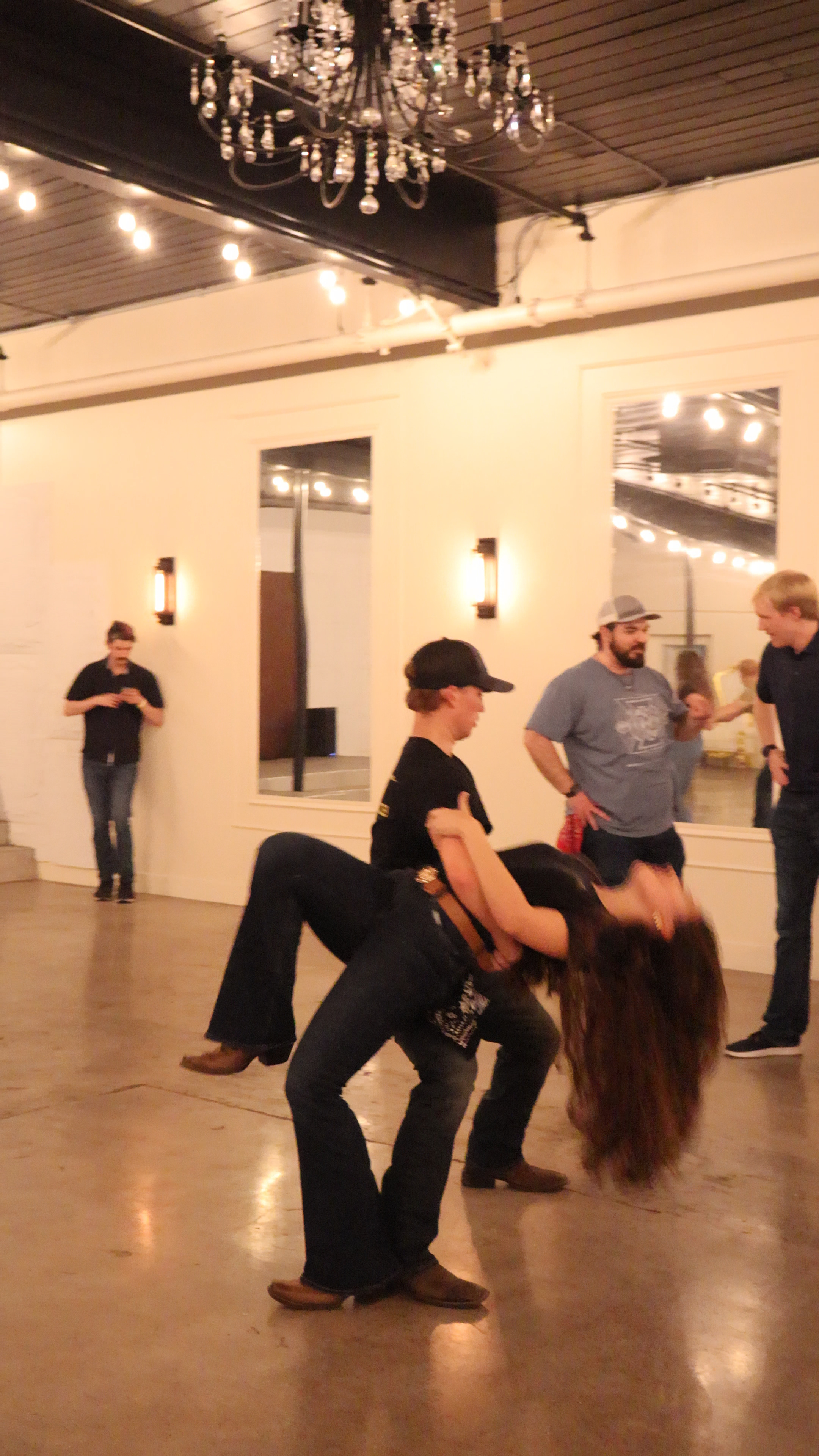 People dancing at a social gathering or party in an indoor venue with warm lighting, chandeliers, and large mirrors on the wall.